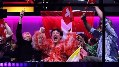 Der Eurovision Song Contest 2025 wird in Basel ausgetragen. Nemo, Person mit braunen lockigen Haaren, schwingt begeistert eine Flagge der Schweiz. 