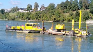 Auf einem Ponton auf dem Wasser heben gelbe Maschinen akutell probeweise ein Stück des Rhein-Flussbetts aus.