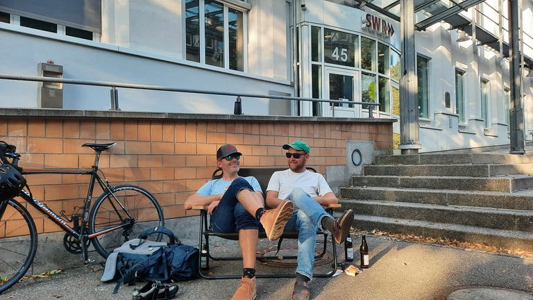 Zwei Männer sitzen auf einer Campingbank vor dem SWR Studio Freiburg und trinken Bier