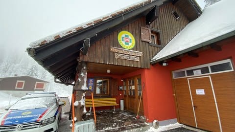 Ein altes kleines Gebäude aus Holz im Schnee. Davor parkt ein Einsatzfahrhzeug der Bergwacht. In dem Haus befand sich bis zum Bau- und Sanierungsbeginn die Rettungswache auf der Feldberg Passhöhe.