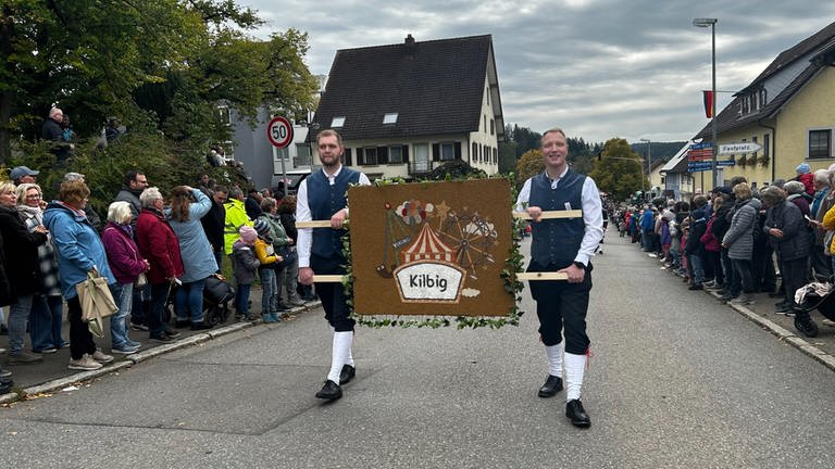 Zwei junge Männer halten ein Bild, das einen Zirkus mit Obst- und Gemüse-Verziehrungen zeigt. Die Männer laufen beim Erntedankumzuzg mit und präsentieren ihr Bild.