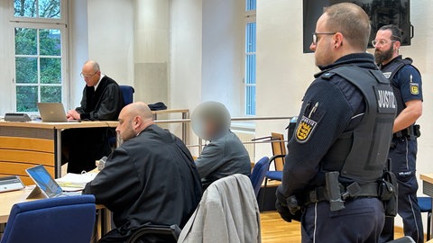 Mann mit grüner Mütze und Schutzmaske sitzt im Gericht. Neben ihm stehen Polizisten. 