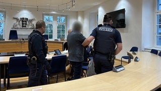 Mann mit grüner Mütze und Schutzmaske sitzt im Gericht. Neben ihm stehen Polizisten. 