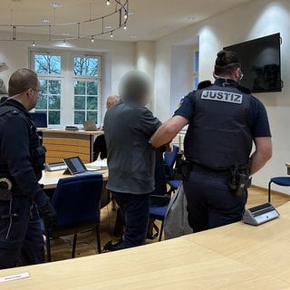 Mann mit grüner Mütze und Schutzmaske sitzt im Gericht. Neben ihm stehen Polizisten. 