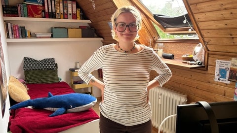 Junge Frau steht in ihrem Mini-WG-Zimmer, das sogar noch Dachschräge hat.