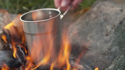 Ein Feuer brennt. Jemand hält einen silbernen Behälter in die Flammen.