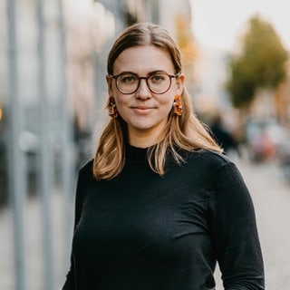 Frau mit Brille und mittellangen Haaren trägt eine Bluse.