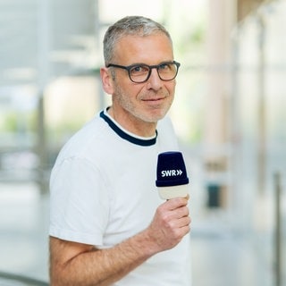 Mann mit Brille und kruzen Haaren lächelt in die Kamera. Er hält ein Mikro mit der Aufschrift "SWR" in den Händen und trägt ein weißes T-Shirt.