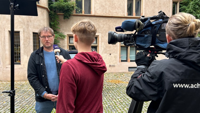 Ein Mann steht vor der Kamera und wird von einem Schüler vor einer Fernsehkamera interviewt.