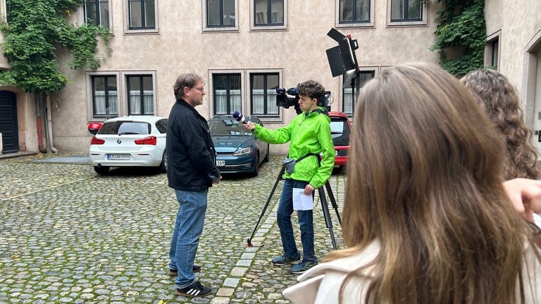 Ein Mann steht vor der Kamera und wird von einem Schüler vor einer Fernsehkamera interviewt.