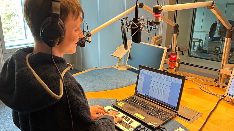 Ein Jugendlicher, der blind ist, steht vor einem Mikrofon im Radiostudio und liest einen Nachrichtentext mithilfe seines Braille-Zeilers vor.