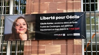 Plakate fordern die freilassung von Cécile Kohler. Neben weißem Text auf schwarzen Grund ist ihr Foto zu sehen.