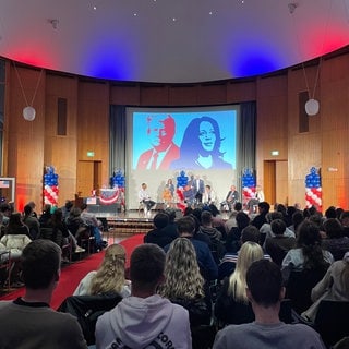 Viele Menschen in der Uni Freiburg schauen auf die Bühne mit der großen Leinwand dahinter. Darauf zu sehen: Donald Trump und Kamala Harris. In der Nacht haben die USA gewählt.