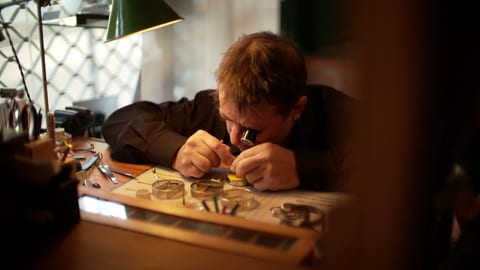 Ein Uhrmachermeister beugt sich an seinem Schreibtisch über eine Uhr, an der er arbeitet.