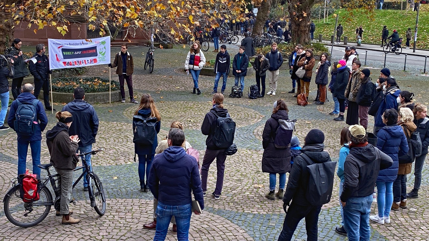 Die Teilnehmenden der Kundgebung stehen im Halbkreis um die Sprechende. Sie spricht in das Mikro und neben ihr halten zwei Organisatioren ein Transparent hoch: 