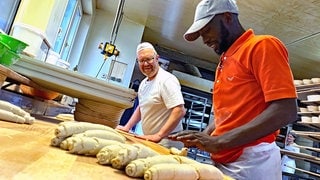 Mit seinen Händen rollt der Bäcker-Lehrling den Teig. Er und sein Lehrmeister strahlen. 