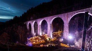 Der Weihnachtsmarkt in der Ravennaschlucht mit der historischen Eisenbahnbrücke erstahlt in der Dunkelheit in leuchtenden Farben. 