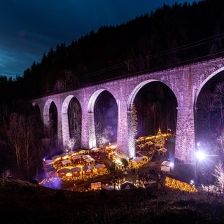 Der Weihnachtsmarkt in der Ravennaschlucht mit der historischen Eisenbahnbrücke erstahlt in der Dunkelheit in leuchtenden Farben. 