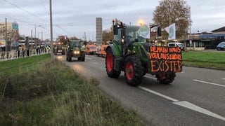 Grüner Traktor mit einem großen Schild fährt über eine Straße, ihm folgen weitere mit französischer Nationalflagge