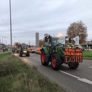 Grüner Traktor mit einem großen Schild fährt über eine Straße, ihm folgen weitere mit französischer Nationalflagge