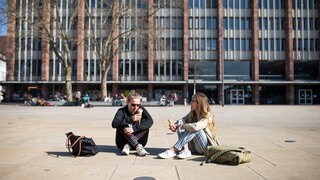 Zwei Studentinnen sitzen mit einem Eis auf dem Platz der alten Synagoge in der Sonne.