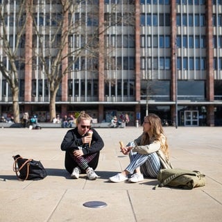 Zwei Studentinnen sitzen mit einem Eis auf dem Platz der alten Synagoge in der Sonne.