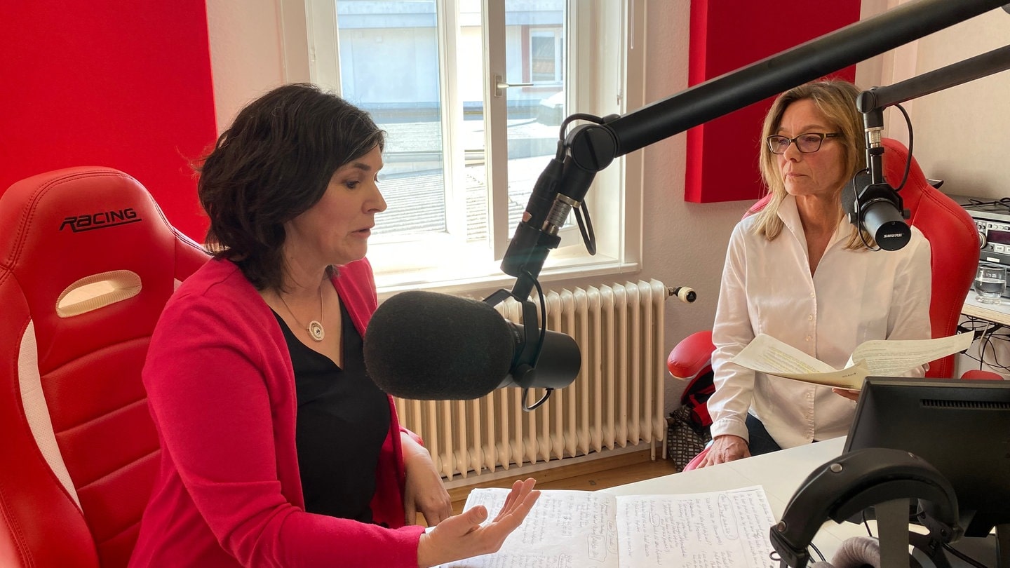 Zwei Frauen sitzen nebeneinander. Die Frau links hat schwarze, kinnlange Haare und trägt einen pinken Blazer. Die Frau rechts hat schulterlange, blonde Haare und trägt eine weiße Bluse. Vor beiden Frauen stehen Mikrofone.