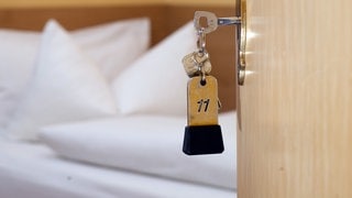 Ein Schlüssel steckt im Schlüsselloch eines Hotelzimmers. Im Hintergrund sieht man ein säuberlich gemachtes Hotelbett. 