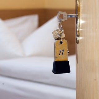 Ein Schlüssel steckt im Schlüsselloch eines Hotelzimmers. Im Hintergrund sieht man ein säuberlich gemachtes Hotelbett. 