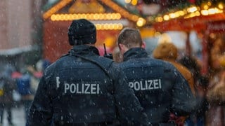 Zwei Polizisten stehen auf einem Weihnachtsmarkt. Man sieht ihren Rücken, die beiden Sprechen miteinander.