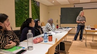 Vier zugewanderte Frauen sitzen an Tischen und hören einer Lehrerin zu. Die Lehrerin steht vor einer Tafel, auf der steht: Feste feiern. Auf den Tischen sind Lehrbücher, Blöcke und Trinkflaschen. Es ist ein Eltern-Integrationskurs in Freiburg-Weingarten.