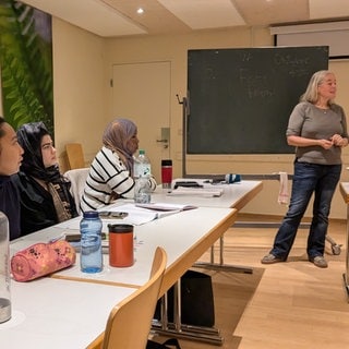 Vier zugewanderte Frauen sitzen an Tischen und hören einer Lehrerin zu. Die Lehrerin steht vor einer Tafel, auf der steht: Feste feiern. Auf den Tischen sind Lehrbücher, Blöcke und Trinkflaschen. Es ist ein Eltern-Integrationskurs in Freiburg-Weingarten.