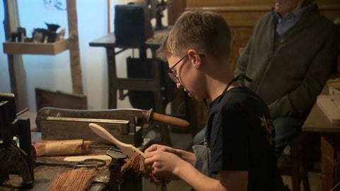 Der 13-jährige Felix sitzt an einer Werkbank und fertigt eine Bürste an. Das Handwerk hat er vom Opa gelernt.