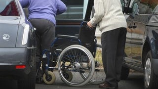 Eine gehbehinderte Frau steigt aus ihrem Rollstuhl ins Auto, ein Mann hilft ihr dabei