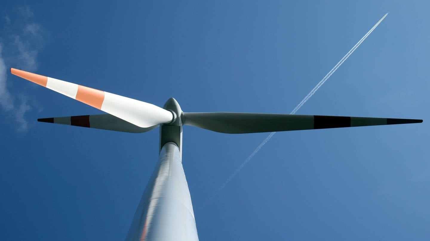 Eine Windkraftanlage von unten vor einem blauen Himmel. Dort fliegt ein Flugzeug in Richtung der rechten, oberen Bildecke.