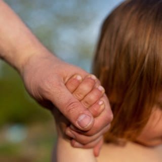 Eine Männerhand hält eine Kinderhand, unscharf dahinter ein kleines Kind mit langen Haaren erkennbar. 