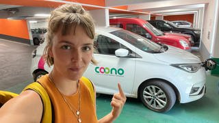 Reporterin Theresa Krampfl steht vor einem E-Auto des Carsharing-Anbieters Coono. 