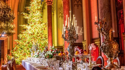 Festlich gedeckte Tafel mit geschmücktem Weihnachtsbaum im Hintergrund und prunkvollen Kerzenständern