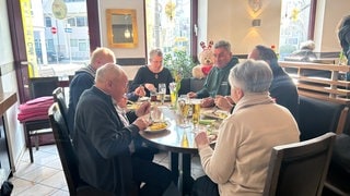 Man sieht einen runden Tisch mit zwei Frauen und vier Männern. Sie essen in einem Restaurant. Auf dem Tisch steht Essen und Trinken.