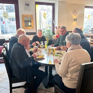 Man sieht einen runden Tisch mit zwei Frauen und vier Männern. Sie essen in einem Restaurant. Auf dem Tisch steht Essen und Trinken.