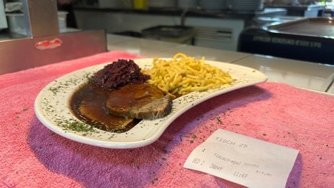 Man sieht einen Teller. Auf dem Teller ist Essen: Schweinebraten mit Rotkohl und Spätzle. Daneben liegt ein Bon aus der Kasse. Auf ihm steht: "Fleisch egal".