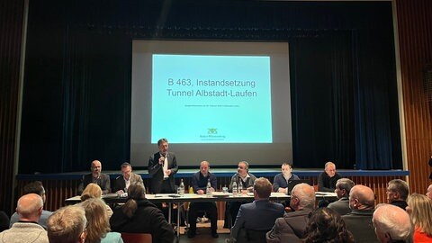 Zu sehen ist die Informationsveranstaltung zur Tunnelsanierung auf der B463. Albstadts Oberbürgermeister Roland Tralmer spricht vor den Zuhörerinnen und Zuhörern.