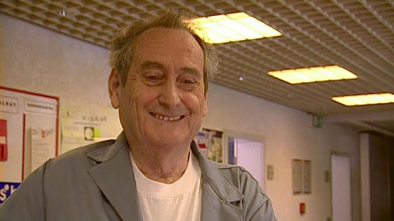 Walter Schultheiß lächelt in grauem Hausmeisterkittel, bekleidet mit einem weissen SWR-Shirt in die Kamera