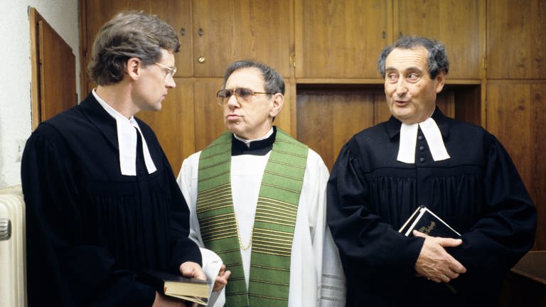 Walter Schultheiß rechts im Bild als Pfarrer verkleidet mit Bibel in der Hand schaut nach links auf zwei weitere Pfarrerkollegen. Ein Ausschnitt aus der Serie: "Oh Gott, Herr Pfarrer".