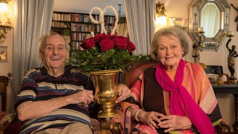 Walter Schultheiß hält in der Hand einen goldfarbenen Weinkühler mit roten Rosen, über denen in weiß die Uahl 90 prangt. Den Strauß gab es für seine rechts neben ihm im Bild sitzende Ehefrau Trudel Wulle. 