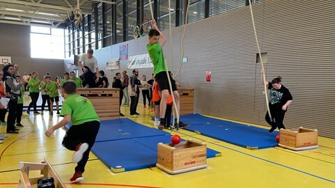 Ein Jugendlicher schwingt an einem Seil in einer Turnhalle, ein anderer rennt mit einem Ball zurück zur Staffelübergabe