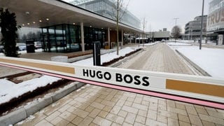 Man sieht eine Schranke. Auf dieser steht: "Hugo Boss". Dahinter sieht man mehrere Gebäude entlang einer langen Straße.