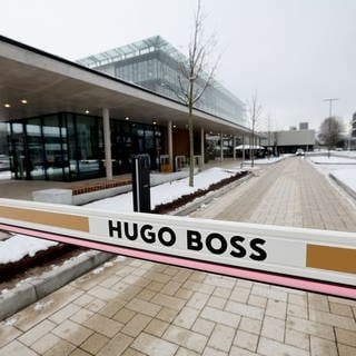 Man sieht eine Schranke. Auf dieser steht: "Hugo Boss". Dahinter sieht man mehrere Gebäude entlang einer langen Straße.