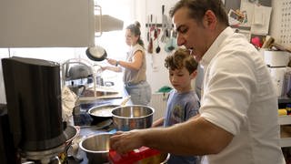 Alejandro Cardosa Zea mit seinem Sohn Leo in der Küche am Herd. Im Hintergrund die Mutter Anna Markovic. Die Eltern haben sich bei einem Erasmus Semester kennengelernt.
