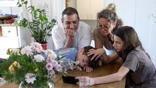 Die Familie Cardosa Markovic sitzt an einem Tisch und spricht über das Handy mit der Oma in Sevilla. Die Eltern haben sich bei einem Erasmussemester kennengelernt.